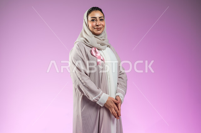شعار اليوم العالمي للتوعيه بالكشف المبكر لسرطان الثدي، الحملة التوعويه بالامراض الخبيثه، بورتريه لامرأة عربية خليجية سعودية مبتسمة محجبة ترتدي عباءه مع الشريط الوردي كفيها متلامسان باتجاه الاسفل، خلفية ورديه، محتوى حساس، استخدام حساس