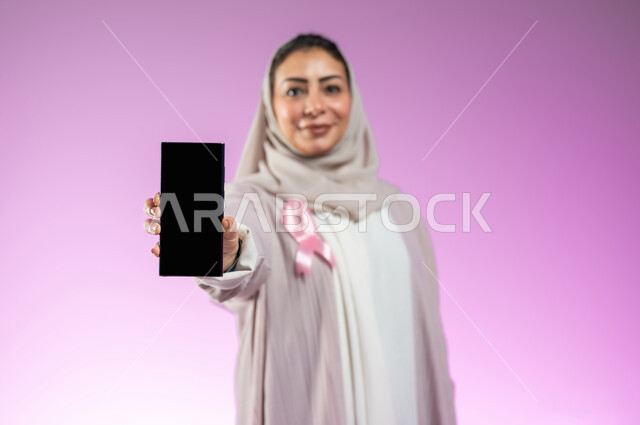 شعار اليوم العالمي للتوعيه بالكشف المبكر لسرطان الثدي، الحملة التوعويه بالامراض الخبيثه، بورتريه لشابة عربية خليجية سعودية مبتسمة محجبة ترتدي العباءة مع الشريط الوردي تقوم بعرض شاشة فارغة سوداءعبر الهاتف المحمول، خلفية ورديه، محتوى حساس، استخدام حساس