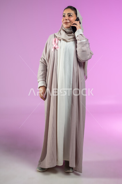 شعار اليوم العالمي للتوعيه بالكشف المبكر لسرطان الثدي، الحملة التوعويه بالامراض الخبيثه، النظر الى الافق البعيدة بنظرات مستقبلية، بوتريه لامرأة عربية خليجية سعودية مبتسمة محجبة ترتدي العباءه مع شريط وردي تتحدث عبر الهاتف المحمول، صورة الجسم بالطول الكامل، خلفية ورديه، محتوى حساس، استخدام حساس