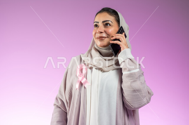 The logo of the global campaign to challenge malignant diseases, a portrait of a smiling Saudi Arabian Gulf woman, veiled and wearing an abaya, talking on a mobile phone, pink background, sensitive content, sensitive use