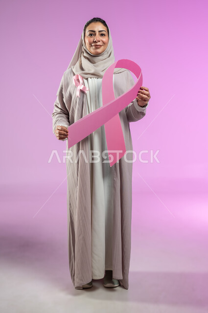 شعار الحملة العالمية لتحدي الأمراض الخبيثة، بورتريه لامرأة عربية خليجية سعودية مبتسمة محجبة ترتدي العباءة تحمل شعار الحملة ضد سرطان الثدي، صورة الجسم بالطول الكامل، خلفية ورديه، محتوى حساس، استخدام حساس