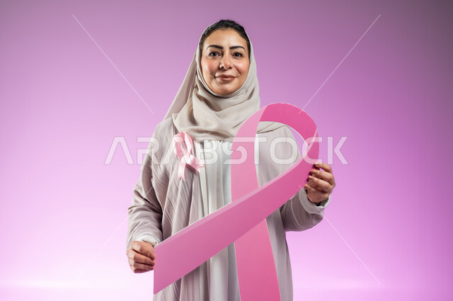 الحملة التوعويه بالامراض الخبيثه، بورتريه مقرب لشابة عربية خليجية سعودية محجبة مبتسمة ترتدي العباءة الملونة مع الشريط الوردي تمسك بيديها شعار اليوم العالمي للتوعيه بالكشف المبكر لسرطان الثدي، خلفية ورديه, محتوى حساس, استخدام حساس