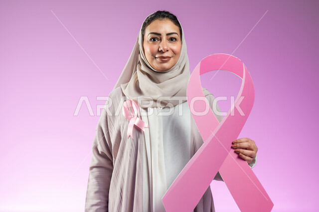 الحملة التوعويه بالامراض الخبيثه،بورتريه مقرب لشابة عربية خليجية سعودية محجبة مبتسمة ترتدي العباءة الملونة مع الشريط الوردي تمسك بيدها شعار اليوم العالمي للتوعيه بالكشف المبكر لسرطان الثدي، خلفية ورديه, محتوى حساس, استخدام حساس