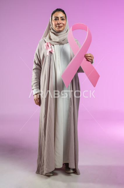 الحملة التوعويه بالامراض الخبيثه، بورتريه لامرأة عربية خليجية سعودية مبتسمة محجبة ترتدي العباءة مع الشريط الوردي  تحمل شعار اليوم العالمي للتوعيه بالكشف المبكر لسرطان الثدي، صورة الجسم بالطول الكامل، خلفية ورديه، محتوى حساس، استخدام حساس