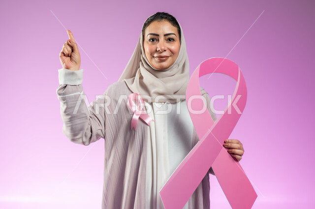 الحملة التوعويه بالامراض الخبيثه، الأشارة إلى شيء ما في الأعلى، بورتريه مقرب لامرأة عربية خليجية سعودية مبتسمة محجبة ترتدي العباءة مع الشريط الوردي تحمل شعار اليوم العالمي للتوعيه بالكشف المبكر لسرطان الثدي، خلفية ورديه، محتوى حساس، استخدام حساس