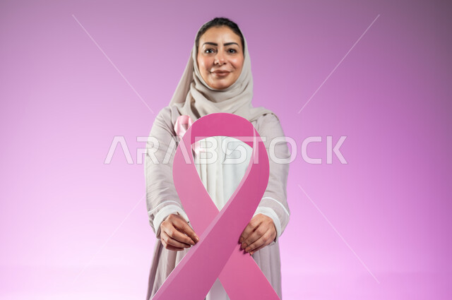 الحملة التوعيه بالامراض الخبيثة, بورتريه مقرب لشابة عربية خليجية سعودية محجبة مبتسمة ترتدي العباءة الملونة تمسك بيدهاشعار التوعيه للكشف المبكر لسرطان الثدي، خلفية ورديه, محتوى حساس, استخدام حساس