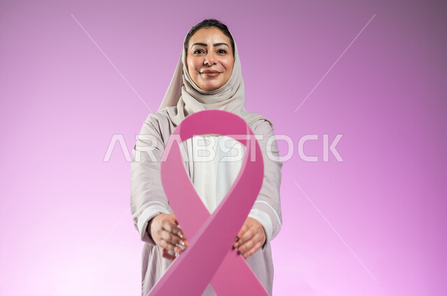 الحملة التوعويه بالامراض الخبيثه، بورتريه مقرب لشابة عربية خليجية سعودية محجبة مبتسمة ترتدي العباءة الملونة تمسك بيدها شعار التوعيه بالكشف المبكر لسرطان الثدي, خلفية ورديه, محتوى حساس, استخدام حساس
