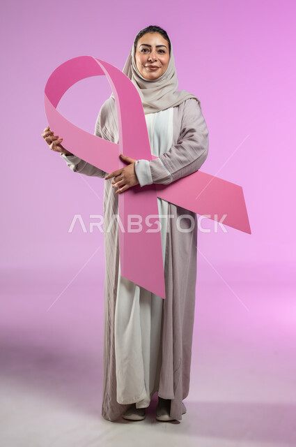 الحملة التوعيه بالامراض الخبيثة, بورتريه مقرب لشابة عربية خليجية سعودية محجبة مبتسمة ترتدي العباءة الملونة تمسك بيدها شعار التوعيه بالكشف المبكر لسرطان الثدي, صورة االجسم بالطول الكامل، خلفية ورديه, محتوى حساس, استخدام حساس