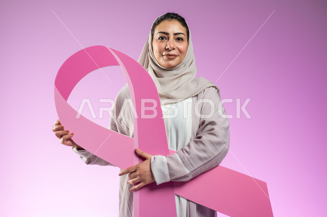 الحملة التوعيه بالامراض الخبيثة, بورتريه مقرب لشابة عربية خليجية سعودية محجبة مبتسمة ترتدي العباءة الملونة تمسك بيدها شعار الحملة العالمية للكشف المبكر عن سرطان الثدي, خلفية ورديه, محتوى حساس, استخدام حساس