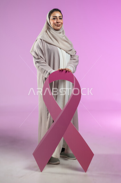 النظر الى الكاميرا بتعابير القوة، بورتريه لامرأة عربية خليجية سعودية مبتسمة محجبة ترتدي العباءة تمسك شعار اليوم العالمي للتوعيه بالكشف المبكر لسرطان الثدي، الحملة التوعويه بالامراض الخبيثه، صورة الجسم بالطول الكامل، خلفية ورديه، محتوى حساس، استخدام حساس