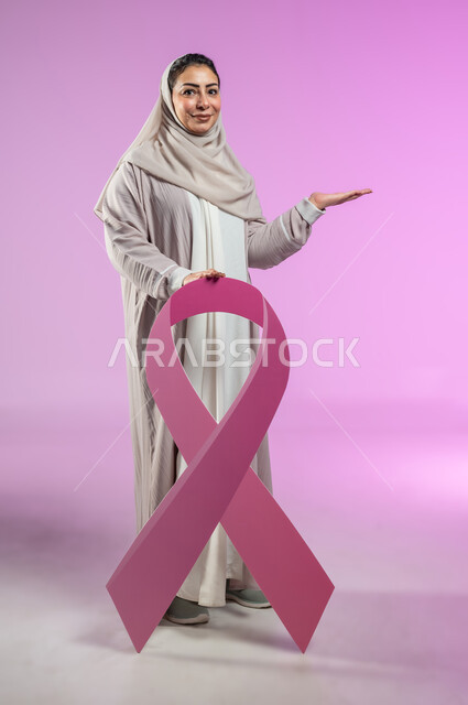 شعار اليوم العالمي للتوعيه بالكشف المبكر لسرطان الثدي، الحملة التوعويه بالامراض الخبيثه، بورتريه لامرأة عربية خليجية سعودية مبتسمة محجبة ترتدي العباءة تمسك بيدها شعار حملة التوعية، الاشارة بالكف إلى شيء ما و حمل عنصر معين، صورة الجسم بالطول الكامل، خلفية ورديه، محتوى حساس، استخدام حساس