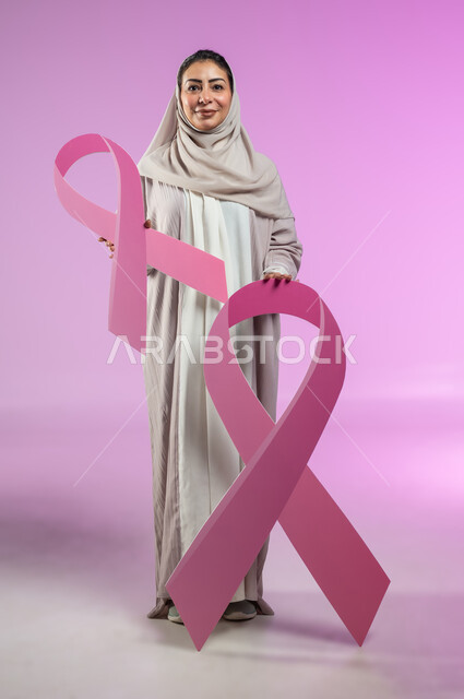 الحملة التوعيه بالامراض الخبيثة, بورتريه لإمرأة عربية خليجية سعودية محجبة مبتسمة ترتدي العباءة الملونة تمسك بيدها شعار التوعيه بالكشف المبكر لسرطان الثدي، النظر إلى الكاميرا بتعابير القوة, صورة الجسم بالطول الكامل, خلفية ورديه, محتوى حساس, استخدام حساس
