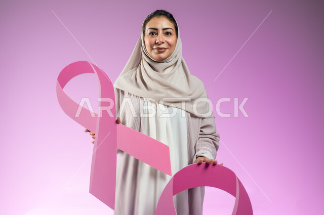 تحدي الأمراض الخبيثة, القيام بحملة توعوية عن سرطان الثدي, النظر إلى الكاميرا بإيماءات السرور, بورتريه مقرب لإمرأة عربية خليجية سعودية ترتدي العباءة الملونة تمسك بيديها شعاري الحملة العالمية، خلفية ورديه, محتوى حساس, استخدام حساس