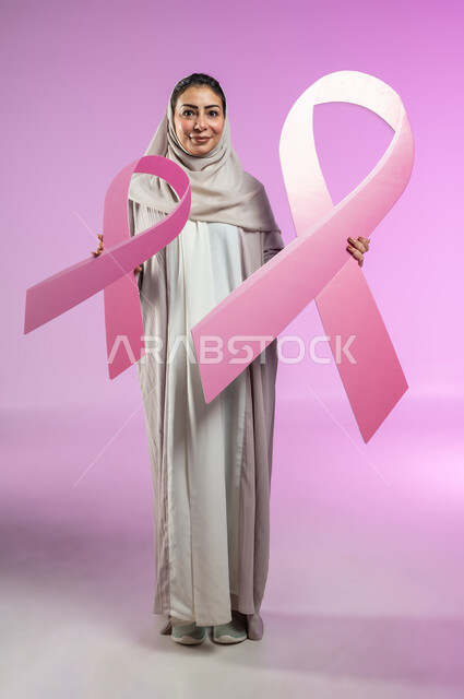 تحدي الأمراض الخبيثة, القيام بحملة توعوية عن سرطان الثدي, النظر إلى الكاميرا بإيماءات السرور, بورتريه لإمرأة عربية خليجية سعودية ترتدي العباءة الملونة تمسك بيديها شعاري الحملة العالمية، الجسم بالطول الكامل، خلفية ورديه, محتوى حساس, استخدام حساس
