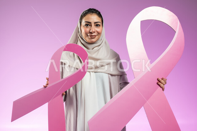 القيام بحملة توعوية عن سرطان الثدي, النظر إلى الكاميرا بإيماءات السرور, تحدي الأمراض الخبيثة, بورتريه مقرب لإمرأة عربية خليجية سعودية ترتدي العباءة الملونة تمسك بيديها شعاري الحملة العالمية، خلفية ورديه, محتوى حساس, استخدام حساس