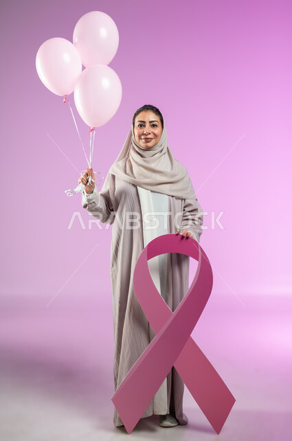 World Breast Cancer Awareness and Early Detection Day, standing confidently and looking at the camera with happy gestures, portrait of a Saudi Arabian Gulf woman wearing an abaya, holding pink balloons in her hand and the global campaign logo, full-length body, pink background, sensitive content, sensitive use