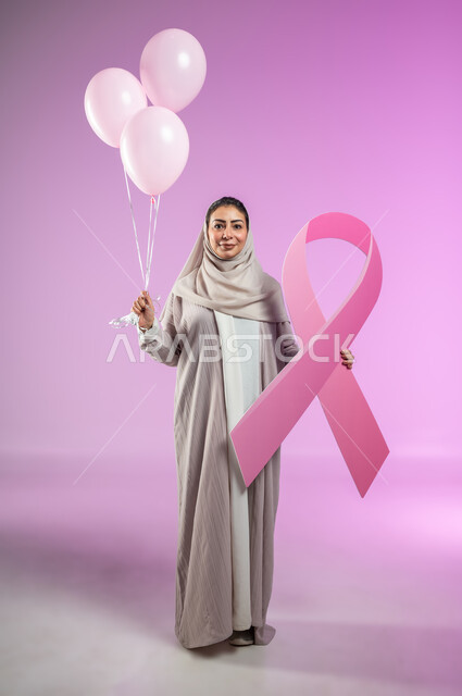 اليوم العالمي للتوعية والكشف المبكر عن سرطان الثدي, النظر إلى الكاميرا بإيماءات السرور, بورتريه لإمرأة عربية خليجية سعودية  ترتدي العباءة تمسك بيدها البلالين الوردية وشعار الحملة العالمية، الجسم بالطول الكامل, خلفية ورديه, محتوى حساس, استخدام حساس