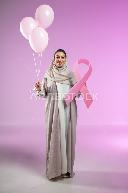 Looking at the camera with gestures of pleasure, World Breast Cancer Awareness and Early Detection Day, portrait of a Saudi Gulf Arab woman wearing an abaya, holding pink balloons in her hand and the global campaign logo, full-length body, pink background, sensitive content, sensitive use