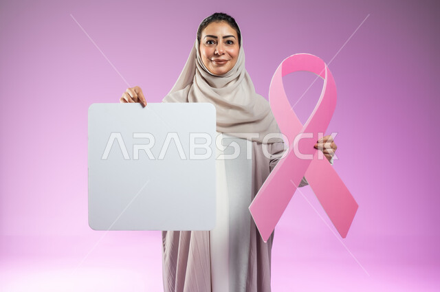لوحات إعلانية للتوعية بسرطان الثدي, بورتريه مقرب لإمرأة عربية خليجية سعودية محجبة مبتسمة ترتدي العباءة تمسك بيديها شعار الحملة العالمية لتحدي الأمراض الخبيثة و موك أب مربع ابيض, خلفية ورديه , محتوى حساس, استخدام حساس