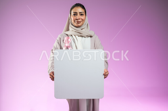 النظر إلى الكاميرا، موك أب أبيض فارغ مربع الشكل، شعار الحملة العالمية لتحدي الأمراض الخبيثة، لوحة بيضاء إعلانية فارغة للتوعية بسرطان الثدي, بورتريه مقرب لامرأة عربية خليجية سعودية مبتسمة محجبة ترتدي العباءة الملونة، خلفية ورديه، محتوى حساس، استخدام حساس