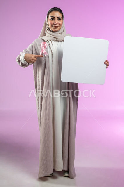 Logo of the global campaign to challenge malignant diseases, billboards for awareness against malignant diseases, portrait of a smiling Saudi Arabian Gulf woman, veiled, wearing an abaya, pointing with her index finger at a blank mockup, full-length body, pink background, sensitive content, sensitive use