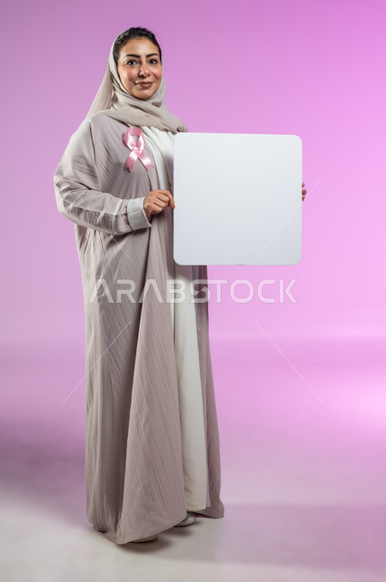 لوحات إعلانية للتوعية ضد الأمراض الخبيثة, النظر إلى الكاميرا بإيماءات السرور، بورتريه لإمرأة عربية خليجية سعودية ترتدي العباءة وتضع شعار الحملة العالمية لسرطان الثدي وتمسك بيدها موك أب مربع فارغ, الجسم بالطول الكامل, خلفية ورديه, محتوى حساس, استخدام حساس