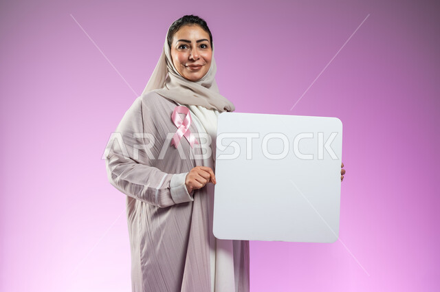 الوقوف و النظر إلى الكاميرا بإيماءات السرور، لوحات إعلانية للتوعية ضد الأمراض الخبيثة,، بورتريه مقرب لإمرأة عربية خليجية سعودية ترتدي العباءة وتضع شعار الحملة العالمية لسرطان الثدي وتمسك بيدها موك أب مربع فارغ, خلفية ورديه, محتوى حساس, استخدام حساس