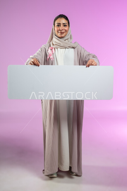 النظر إلى الكاميرا بإيماءات السرور, لوحات إعلانية للتوعية ضد الأمراض الخبيثة, بورتريه لإمرأة عربية خليجية سعودية ترتدي العباءة وتضع شعار الحملة العالمية لسرطان الثدي وتمسك بيدها موك أب مستطيل فارغ, الجسم بالطول الكامل, خلفية ورديه, محتوى حساس, استخدام حساس