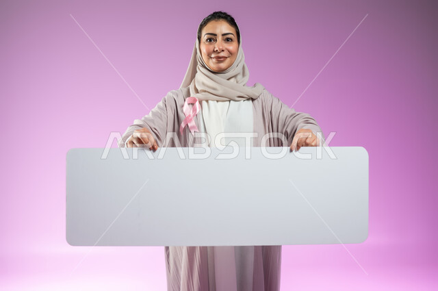 عرض شعار الحملة العالمية لتحدي الأمراض الخبيثة, لوحات إعلانية للتوعية بسرطان الثدي, بورتريه مقرب لإمرأة عربية خليجية سعودية مبتسمة ترتدي العباءة تضع شريط وردي وتمسك بيدها موك أب مستطيل أبيض فارغ , خلفية ورديه, محتوى حساس, استخدام حساس