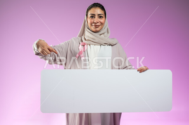 شعار الحملة العالمية لتحدي الأمراض الخبيثة، لوحات إعلانية للتوعية بسرطان الثدي, بورتريه مقرب لامرأة عربية خليجية سعودية مبتسمة محجبة ترتدي العباءة تشير بإصبع السبابة إلى موك أب مستطيل الشكل، خلفية ورديه ، محتوى حساس، استخدام حساس