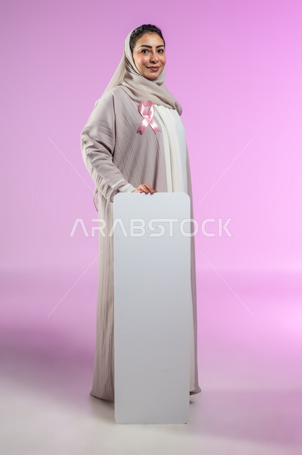 النظر إلى الكاميرا بإيماءات السرور, لوحات إعلانية للتوعية ضد الأمراض الخبيثة, بورتريه لإمرأة عربية خليجية سعودية ترتدي العباءة تضع شعار الحملة العالمية لسرطان الثدي وتمسك بيدها موك أب مستطيل فارغ, الجسم بالطول بالكامل, خلفية ورديه, محتوى حساس, استخدام حساس