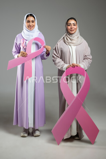 إيماءات السرور والسعادة، اليوم العالمي للكشف عن سرطان الثدي,  بورتريه لامراتان عربيتان خليجيتان سعوديتان محجبتان ترتديان العباءه الملونه تحملان شعار التوعيه للكشف المبكر للأمراض الخبيثة، الجسم بالطول الكامل، خلفية رمادية، محتوى حساس، استخدام حساس