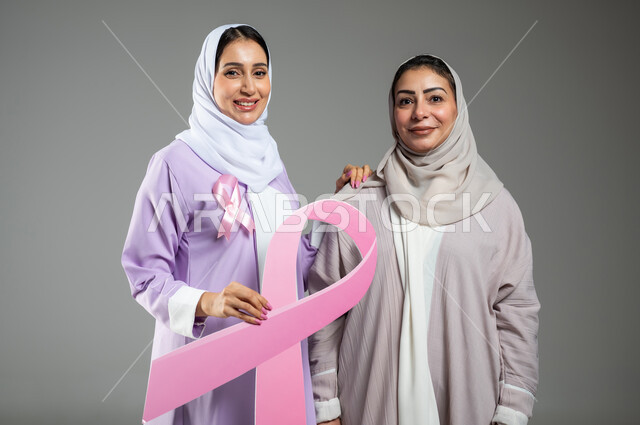 الامساك بشعار التوعيه للكشف المبكر للأمراض الخبيثة، النظر إلى الكاميرا بإيماءات السرور، اليوم العالمي للكشف عن سرطان الثدي,  بورتريه مقرب  لامراتان عربيتان خليجيتان سعوديتان محجبتان ترتديان العباءه الملونه، خلفية رمادية، محتوى حساس، استخدام حساس