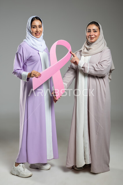النظر إلى الكاميرا بإيماءات السرور, اليوم العالمي للتوعية والكشف المبكر عن سرطان الثدي, بورتريه لامراتان عربيتان خليجيتان سعوديتان محجبتان ترتديان العباءه الملونه، الامساك بشعار الحملة العالمية، الجسم بالطول الكامل، خلفية رمادية، محتوى حساس، استخدام حساس