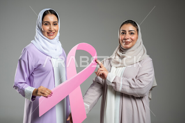 اليوم العالمي للتوعية والكشف المبكر عن سرطان الثدي، النظر إلى الكاميرا بإيماءات السرور, , تحدي الأمراض الخبيثة, بورتريه مقرب لامراتان عربيتان خليجيتان سعوديتان محجبتان ترتديان العباءه الملونه، الامساك بشعار الحملة العالمية، خلفية رمادية، محتوى حساس، استخدام حساس