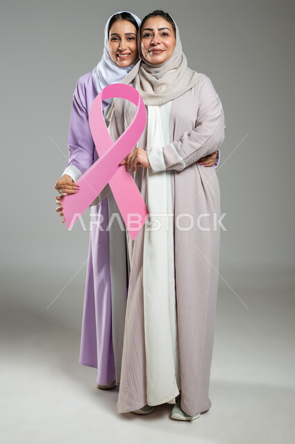 النظر إلى الكاميرا، تحدي الأمراض الخبيثة, اليوم العالمي للكشف عن سرطان الثدي, بورتريه لامراتان عربيتان خليجيتان سعوديتان محجبتان ترتديان العباءه الملونه تمسكان بشعار الحملة العالمية، الجسم بالطول الكامل، خلفية رمادية، محتوى حساس، استخدام حساس