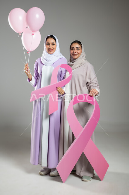 النظر إلى الكاميرا بإيماءات السرور، اليوم العالمي للكشف عن سرطان الثدي،, بورتريه لامراتان عربيتان خليجيتان سعوديتان ترتديان العباءه الملونه تمسكان بشعار التوعيه للكشف المبكر للأمراض الخبيثه، الجسم بالطول الكامل، خلفية رمادية، محتوى حساس، استخدام حساس