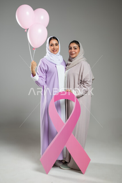 World Breast Cancer Awareness and Early Detection Day, the challenge of malignant diseases, a portrait of two veiled Saudi Gulf Arab women wearing the colorful abaya, holding the global campaign logo and pink balloons, full-length body, gray background, sensitive content, sensitive use