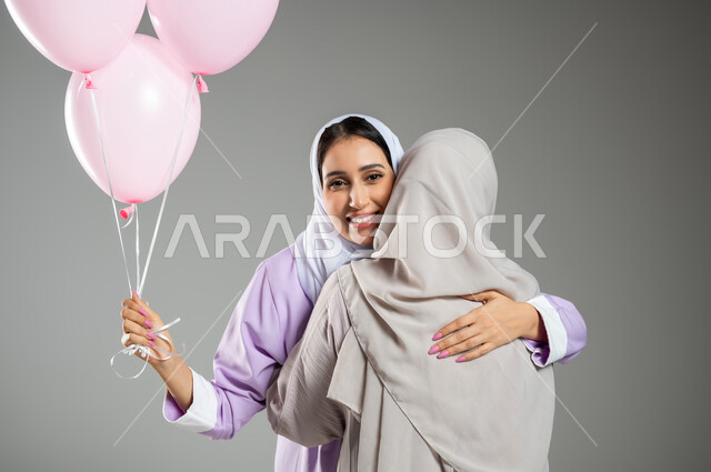 التعاطف والموده بين الاصدقاء، اليوم العالمي للكشف المبكر عن سرطان الثدي, بورتريه مقرب لامراتان عربيتان خليجيتان سعوديتان محجبتان ترتديان العباءه الملونه تقومان بمعانقه بعضهما بحرارة، امساك البلالين الوردية، خلفية رمادية، محتوى حساس، استخدام حساس