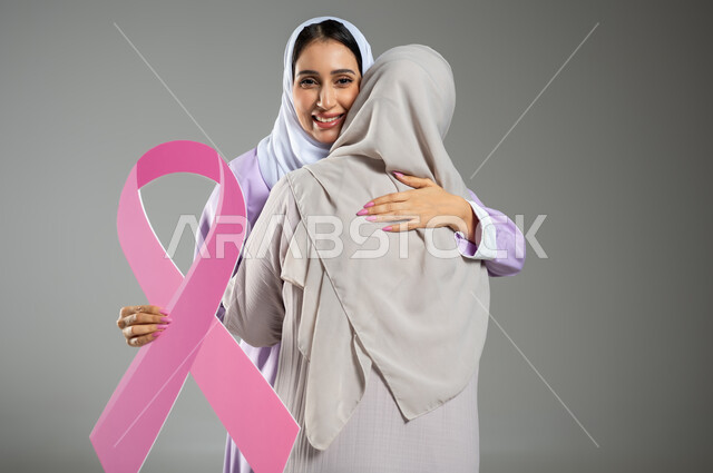 التعاطف والمودة بين الاصدقاء، اليوم العالمي للكشف المبكر عن سرطان الثدي, بورتريه مقرب لامراتان عربيتان خليجيتان سعوديتان محجبتان ترتديان العباءه الملونه تقومان بمعانقه بعضهما بحرارة، خلفية رمادية، محتوى حساس، استخدام حساس