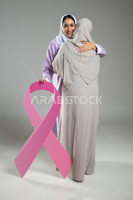 العناق والمودة بين الاصدقاء،الامساك بشعار الحملة العالمية، اليوم العالمي للكشف المبكر عن سرطان الثدي, بورتريه لامراتان عربيتان خليجيتان سعوديتان محجبتان ترتديان العباءه الملونه، الجسم بالطول الكامل، خلفية رمادية، محتوى حساس، استخدام حساس