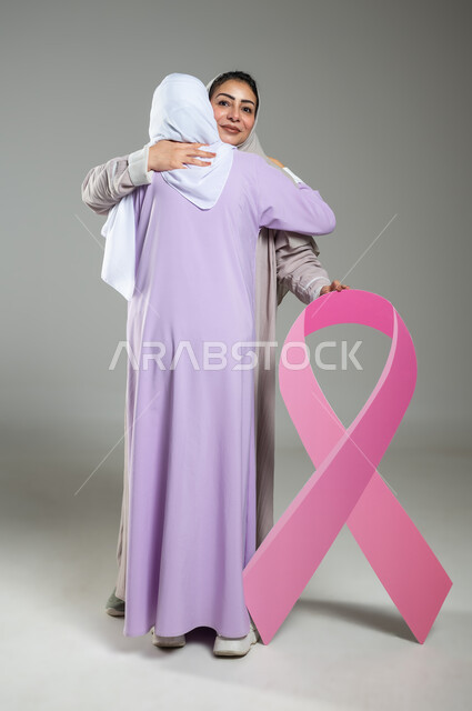 Hugs and affection between friends, holding the global campaign slogan, World Breast Cancer Day, close-up portrait of two veiled Saudi Arabian Gulf women wearing colorful abayas, full-length body, gray background, sensitive content, sensitive use