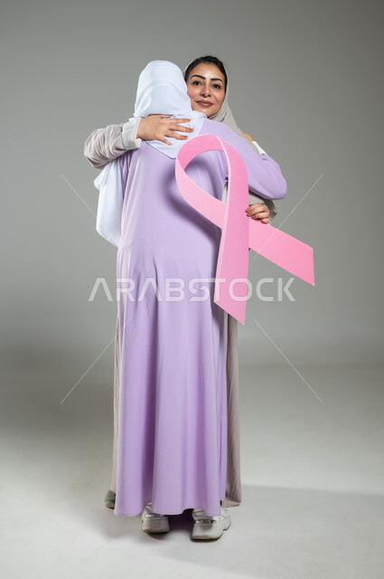 العناق والمودة بين الاصدقاء،الامساك بشعار الحملة العالمية، اليوم العالمي للكشف المبكر عن سرطان الثدي, بورتريه لامراتان عربيتان خليجيتان سعوديتان محجبتان ترتديان العباءه الملونه، الجسم بالطول الكامل، خلفية رمادية، محتوى حساس، استخدام حساس