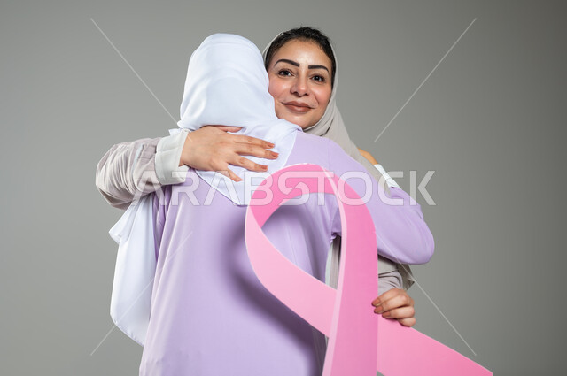 التعاطف والمودة بين الاصدقاء، اليوم العالمي للكشف المبكر عن سرطان الثدي, بورتريه مقرب لامراتان عربيتان خليجيتان سعوديتان محجبتان ترتديان العباءه الملونه تقومان بمعانقه بعضهما بحرارة، خلفية رمادية، محتوى حساس، استخدام حساس