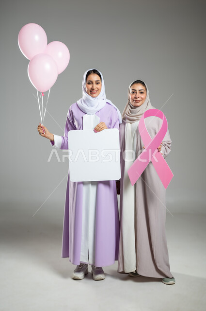 شعار الحملة العالمية لتحدي سرطان الثدي, لوحات إعلانية للتوعية ضد الأمراض الخبيثة، بورتريه لامراتان عربيتان خليجيتان سعوديتان محجبتان ترتديان العباءه الملونه، الامساك بموك أب مربع فارغ وبلالين وردية، الجسم بالطول الكامل, خلفية رمادية، محتوى حساس، استخدام حساس