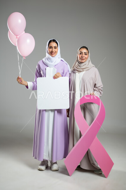 The logo of the global breast cancer challenge campaign, billboards for awareness against malignant diseases, portrait of two veiled Saudi Gulf Arab women wearing colorful abayas, holding an empty square mockup and pink balloons, full length body, gray background, sensitive content, sensitive use