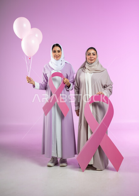 The logo of the Global Campaign to Challenge Breast Cancer, a portrait of two veiled Saudi Arabian Gulf women wearing colorful abayas, holding the Global Campaign logo and pink balloons, full-length body, pink background, sensitive content, sensitive use