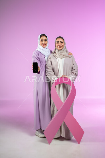 عرض شاشة سوداء فارغة، النظر إلى الكاميرا، شعار الحملة العالمية لتحدي الأمراض الخبيثة، بورتريه لامراتان عربيتان خليجيتان سعوديتان محجبتان ترتديان العباءه الملونه، استخدام الهاتف المحمول، الجسم بالطول الكامل، خلفية ورديه، محتوى حساس، استخدام حساس