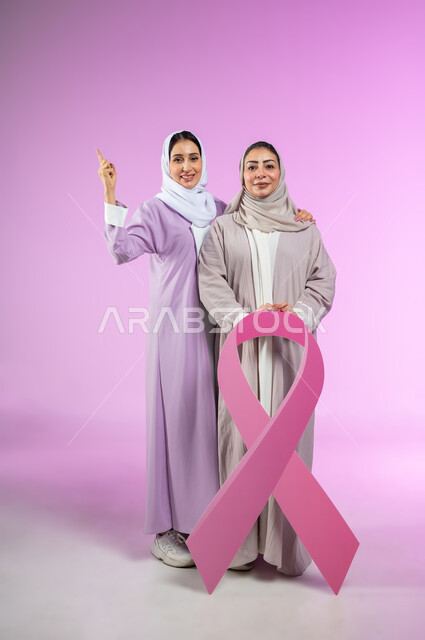 النظر إلى الكاميرا، شعار الحملة العالمية لتحدي الأمراض الخبيثة، بورتريه لامراتان عربيتان خليجيتان سعوديتان محجبتان ترتديان العباءه الملونه، الإشارة بإصبع السبابة إلى الاعلى لوجود شيء ما، الجسم بالطول الكامل، خلفية ورديه، محتوى حساس، استخدام حساس