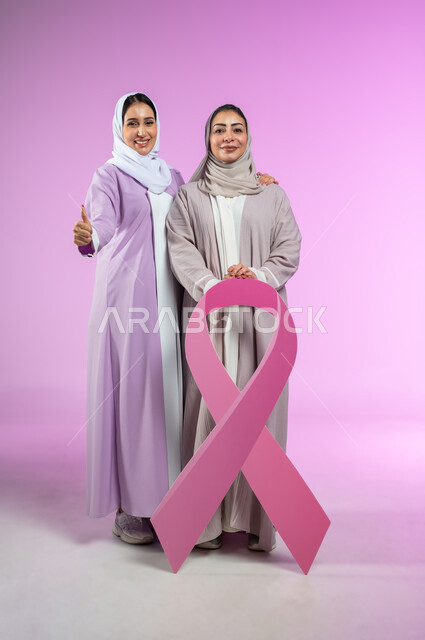 رفع اصبع الابهام بتعابير الرضا والجودة، الاعجاب بشعار الحملة العالمية لتحدي الأمراض الخبيثة، بورتريه لامراتان عربيتان خليجيتان سعوديتان محجبتان ترتديان العباءه الملونه، الجسم بالطول الكامل, خلفية وردية، محتوى حساس، استخدام حساس